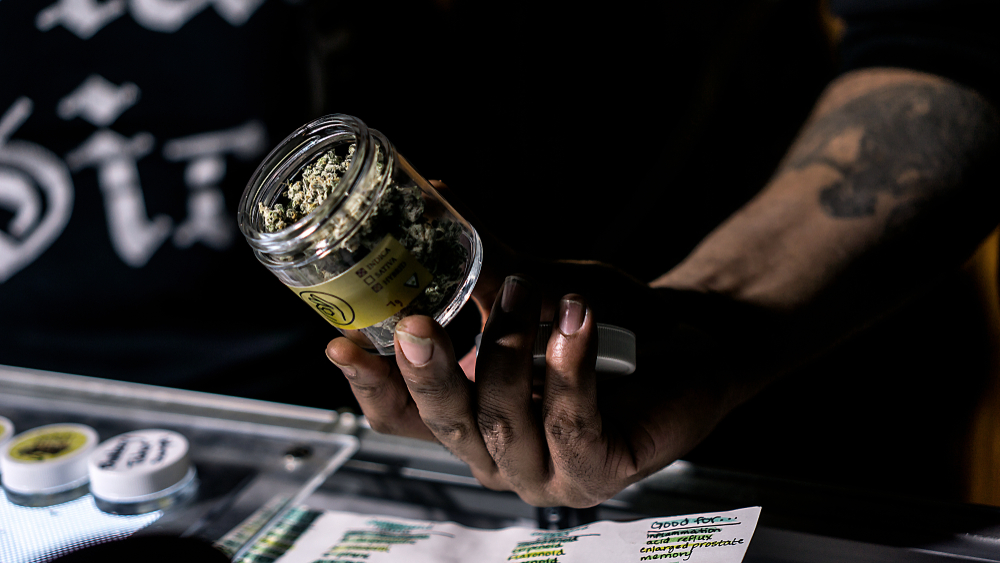 Person holding a jar of cannabis in a dispensary, showcasing the product's contents and labeling, with additional containers visible on the table.