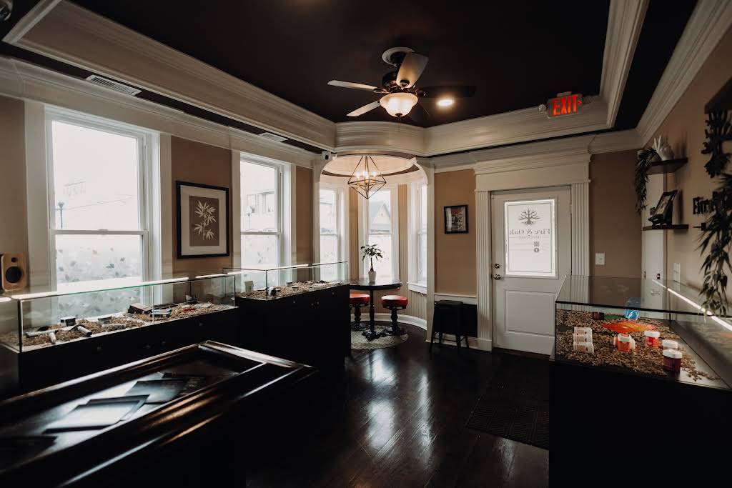 Interior view of a modern cannabis dispensary showcasing display cases filled with products, a stylish seating area, and large windows providing natural light. The space features a contemporary design with dark wood flooring and decorative elements, creating an inviting atmosphere for customers.