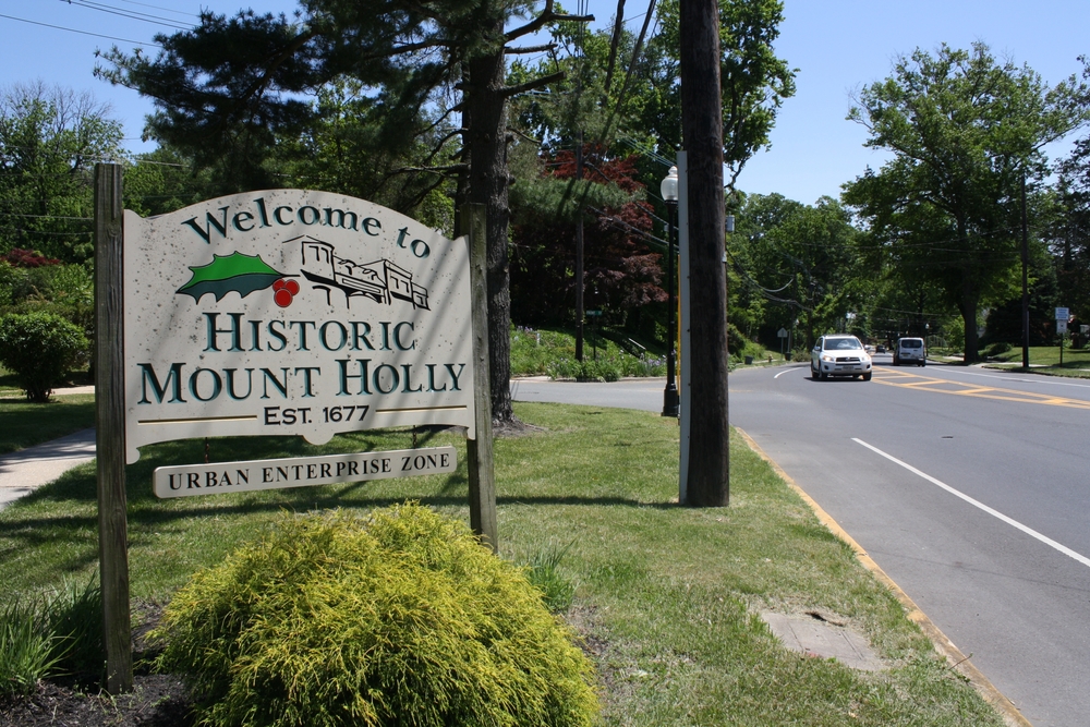 Welcome sign for Historic Mount Holly, established in 1677, located in an urban enterprise zone, with a view of the surrounding greenery and road.